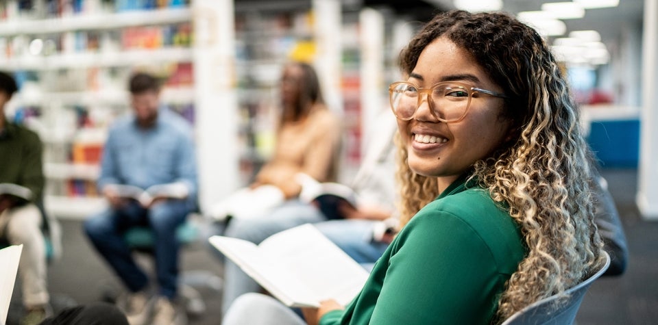 Фотография молодой женщины, сидящей в библиотеке и улыбающейся в камеру.