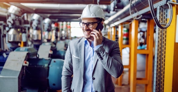 A man in a suit wearing a hardhat speak on his cellphone from inside an industrial area