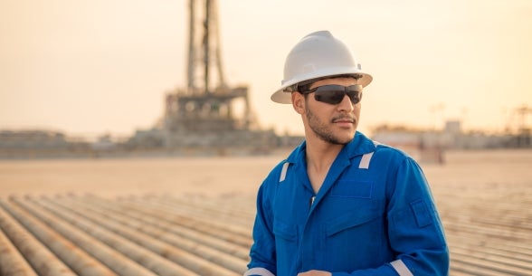 Man in hardhat stands in an oilfield with a rig in the background