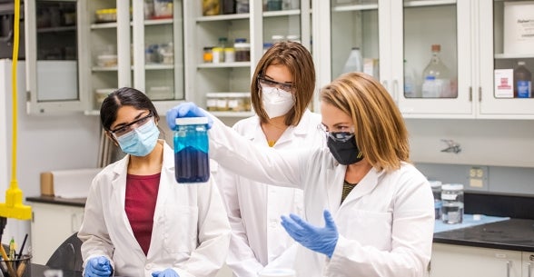 Three students in lab coats, gloves, and masks looking at the contents of a beaker.
