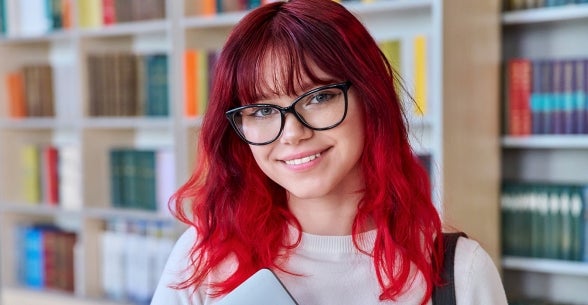 Imagem de uma jovem mulher de óculos em uma biblioteca.