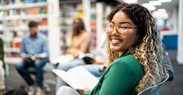 Imagem de uma jovem sentada em uma biblioteca e sorrindo para a câmera.